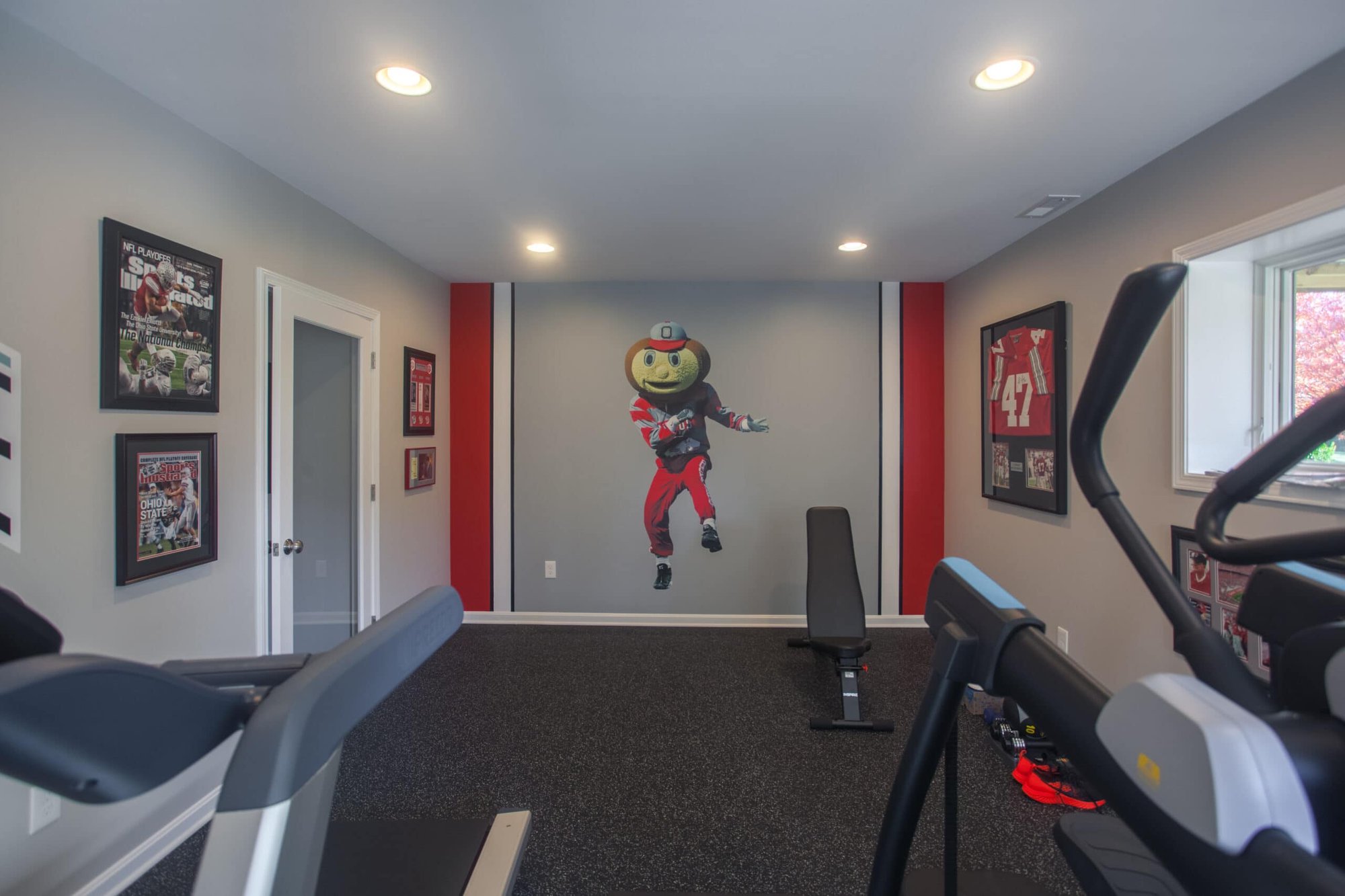 Cincinnati home gym with rubber flooring, workout bench, wall mural, framed sports art, and exercise equipment in a bright space