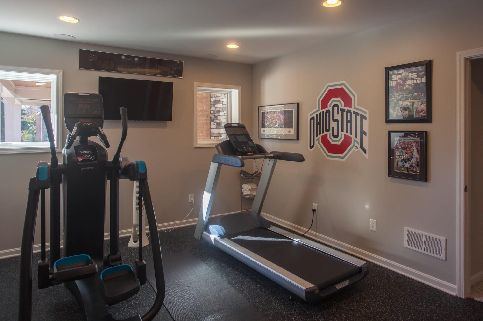 Cincinnati home gym with treadmill, elliptical, wall-mounted TV, Ohio State decor, and rubber flooring in a finished basement space