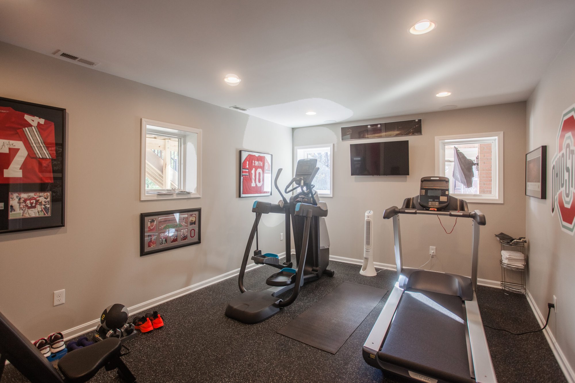 Cincinnati home gym with treadmill, elliptical, wall-mounted TV, framed sports memorabilia, and rubber flooring in a bright finished space