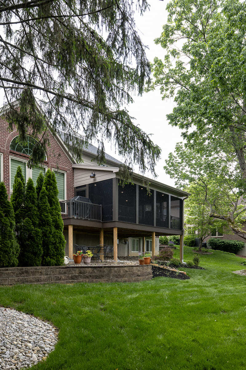 Backyard view of screened deck with wood supports, stairs, lawn, trees, and outdoor patio. Backyard view of screened deck with wood supports, stairs, lawn, trees, and outdoor patio.