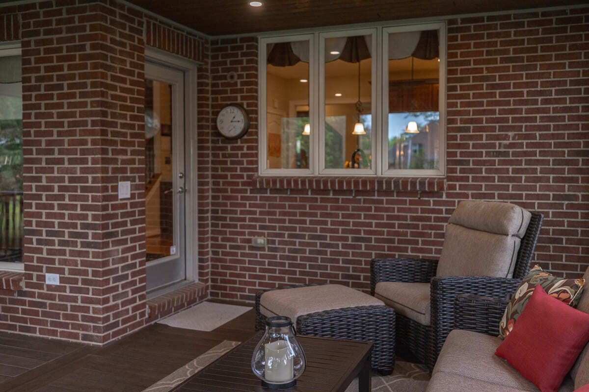 Covered deck lounge with brick wall, wicker chair, wood flooring, and screened outdoor views.