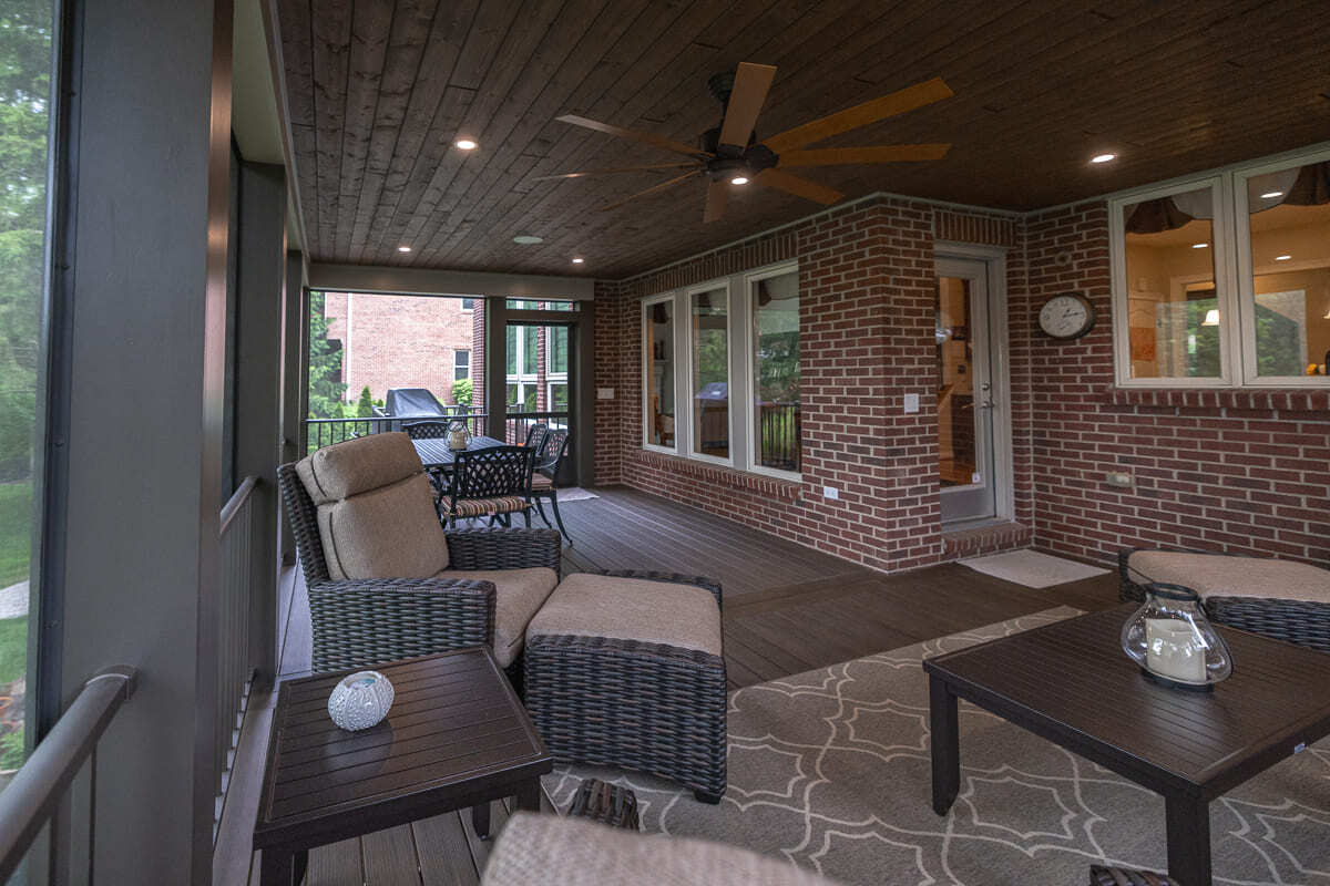 Screened outdoor living space with wood deck flooring, brick wall, fan, and seating areas.