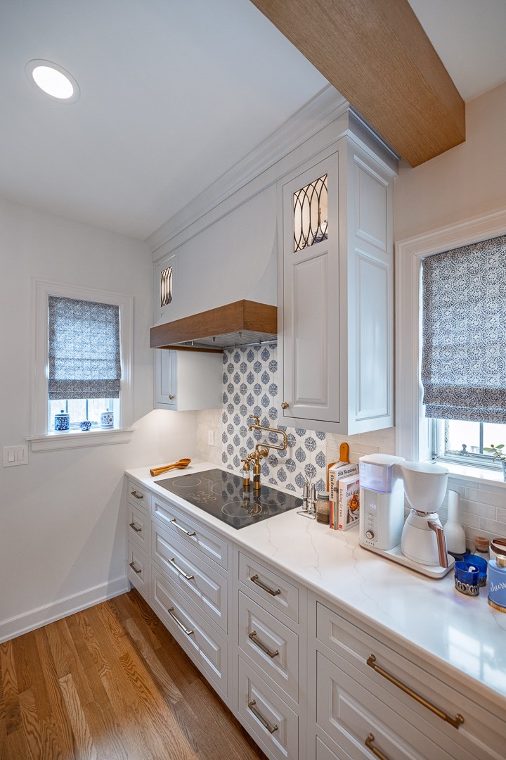 Cincinnati kitchen coffee bar with white cabinetry, brass faucet, patterned backsplash, wood hood accent, and natural light from two windows