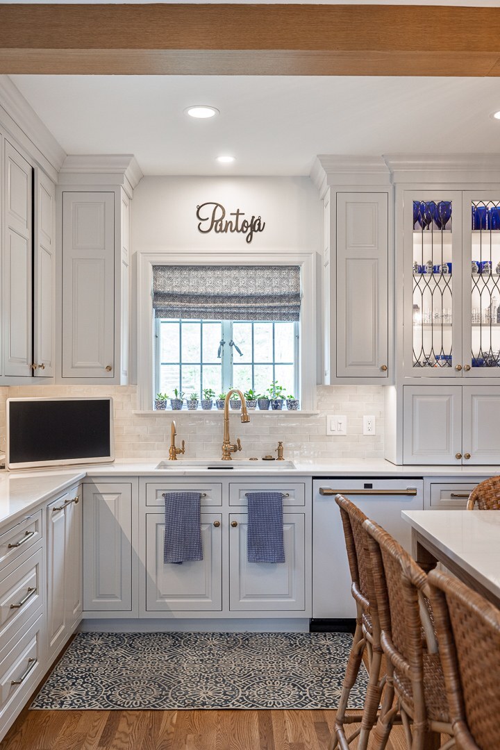 Cincinnati kitchen sink area with white cabinetry, brass faucet, window plants, patterned rug, and glass-front cabinets in a bright space