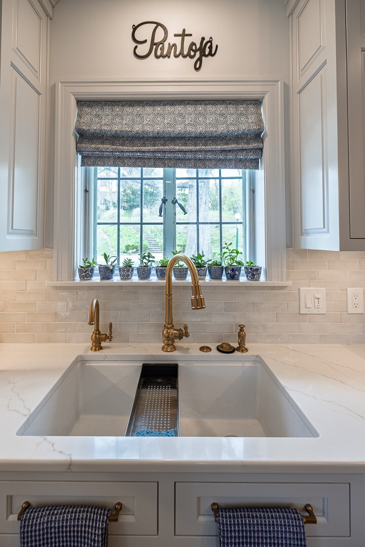 Cincinnati kitchen sink with brass faucet, white farmhouse basin, window with potted plants, and patterned Roman shade in a bright space