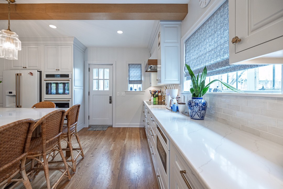 Cincinnati kitchen with white cabinetry, long counter, wood beam ceiling, woven bar stools, and bright natural light from multiple windows Cincinnati kitchen with white cabinetry, long counter, wood beam ceiling, woven bar stools, and bright natural light from multiple windows