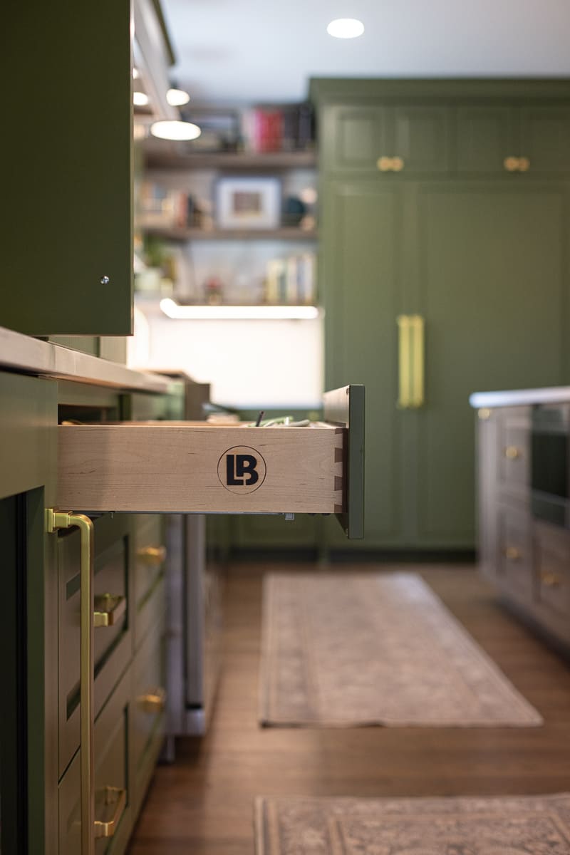 Close-up of custom kitchen drawer with dovetail construction, green cabinetry, brass hardware, and wood flooring. Close-up of custom kitchen drawer with dovetail construction, green cabinetry, brass hardware, and wood flooring.