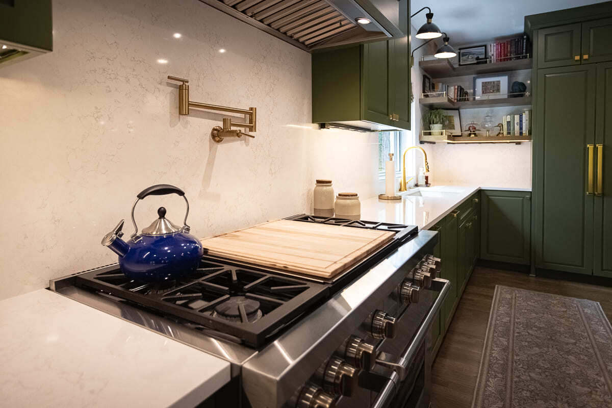 Custom green kitchen cabinetry with stainless steel range, quartz backsplash, brass pot filler, and blue kettle.