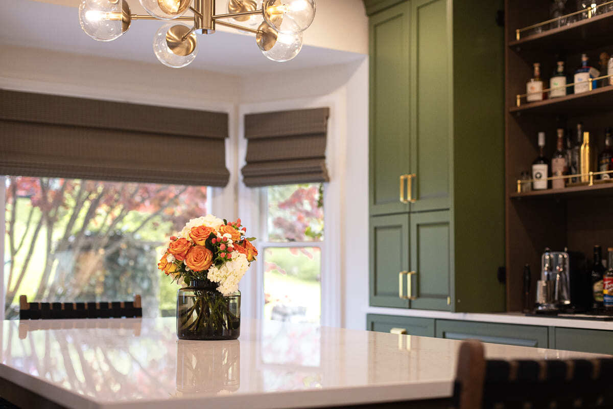 Kitchen dining area with quartz table, green cabinetry, built-in bar shelving, statement lighting, and bay windows.