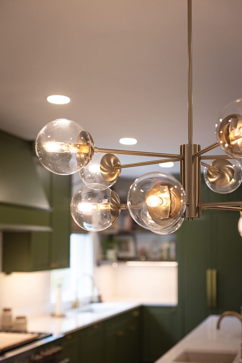 Modern brass chandelier with glass globe lights above a remodeled kitchen with green cabinetry and quartz counters.
