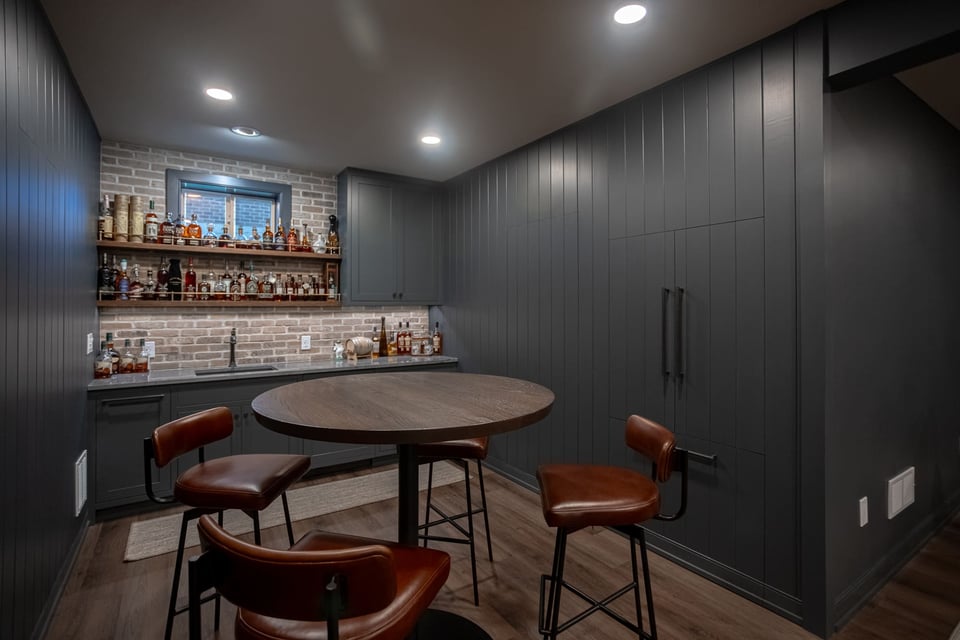 Basement bar with dark cabinetry, wood flooring, brick backsplash, open shelving, and leather seating.