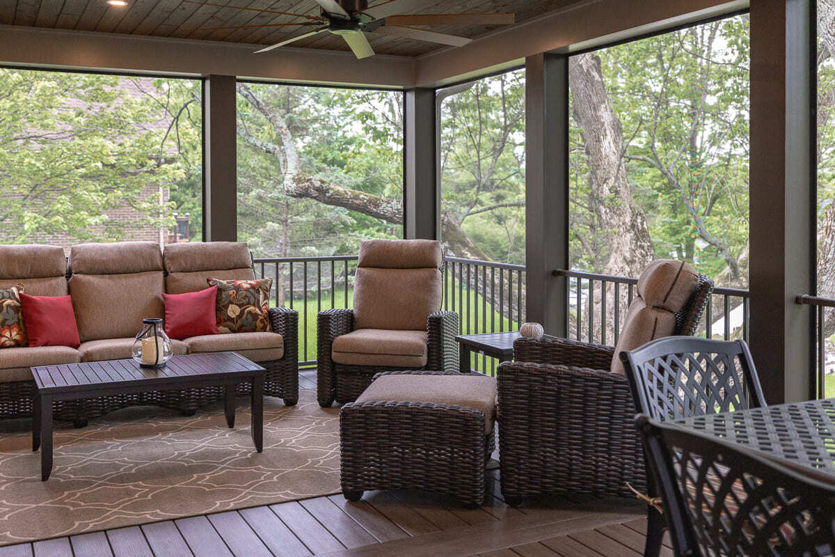 Screened outdoor deck lounge with wicker seating, wood ceiling, railing, and backyard tree views.