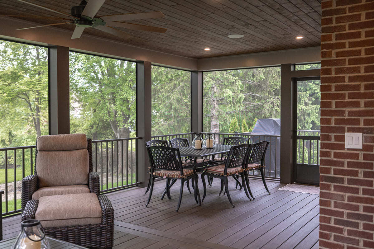 Screened outdoor dining area with wood deck flooring, brick wall, railing, and backyard views.