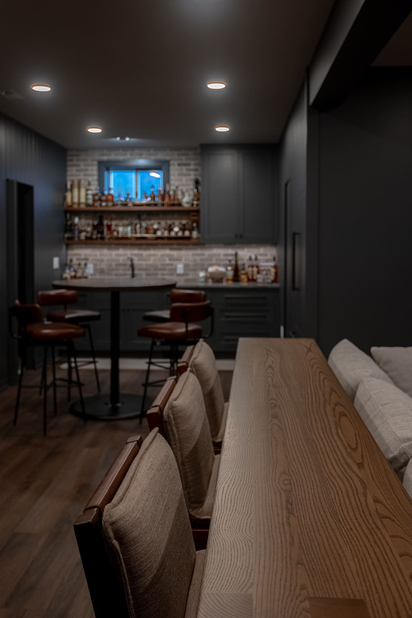 Basement bar and lounge with wood console table, leather seating, dark cabinetry, brick backsplash, and recessed lighting. Basement bar and lounge with wood console table, leather seating, dark cabinetry, brick backsplash, and recessed lighting.